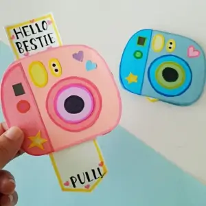 Two finished paper polaroid camera cards, a pink one pulled open reading Hello Bestie and a blue one