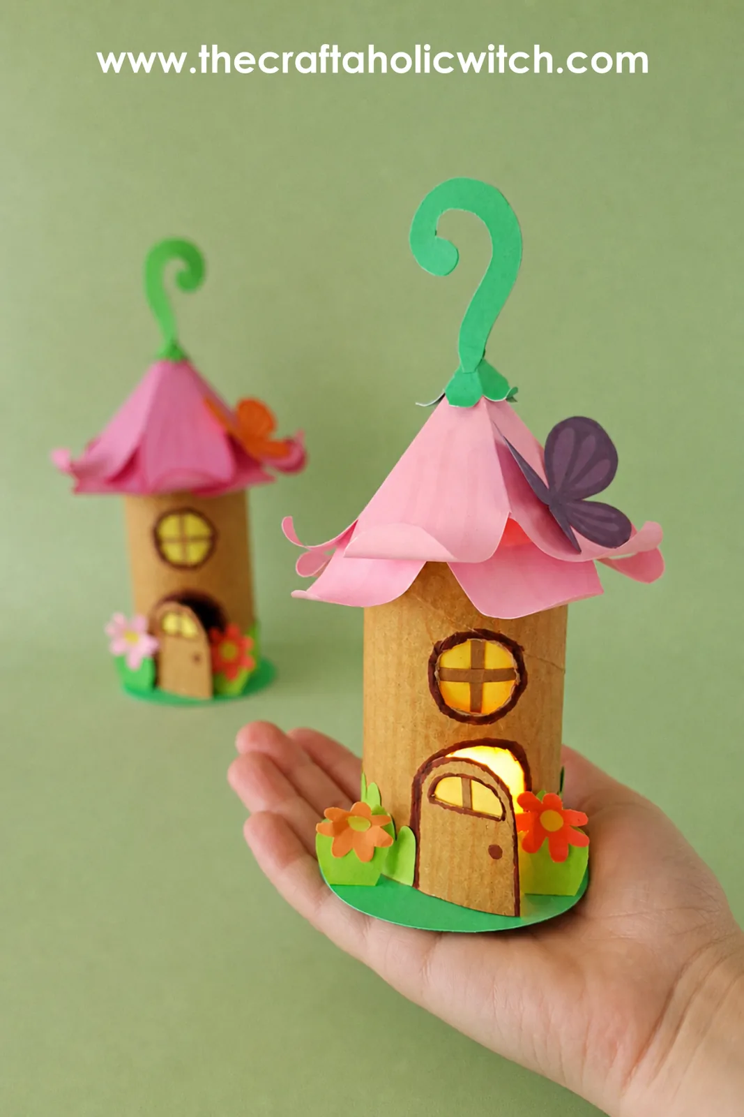 Two toilet paper roll fairy house lanterns with pink flower roofs glowing on a green background