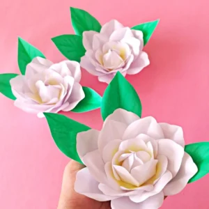 Hand holding a finished realistic paper gardenia flower next to two more blooms and paper leaves