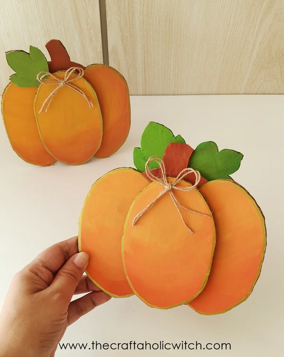 Hand holding a finished cardboard pumpkin craft in front of a second pumpkin