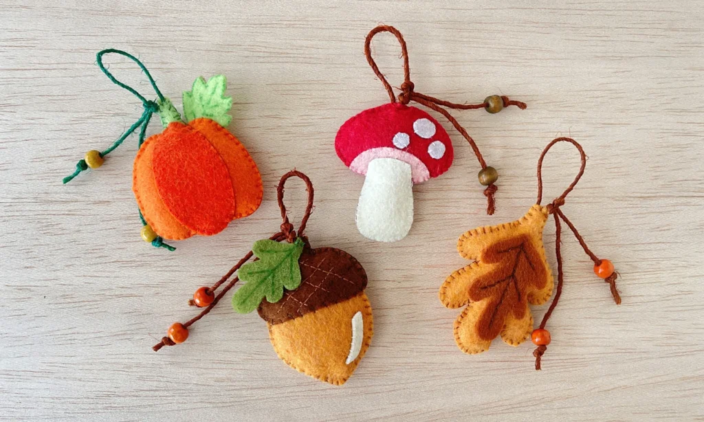Four felt fall ornaments in acorn, mushroom, leaf and pumpkin shapes laid out on a wooden table