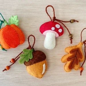 Four felt fall ornaments in acorn, mushroom, leaf and pumpkin shapes laid out on a wooden table