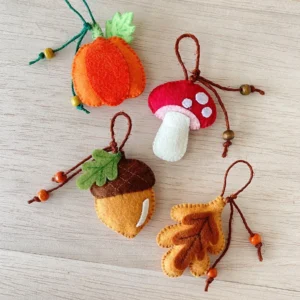 Flat lay of four handmade felt fall ornaments with beaded string loops: a pumpkin, mushroom, acorn and leaf